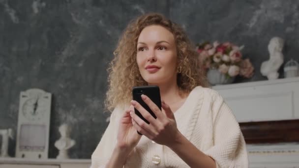 Mladý kavkazský blond kudrnatý žena myslí a píše odpověď na její chytrý telefon záběr ve zpomaleném filmu z nízkého úhlu s květinami na pozadí — Stock video