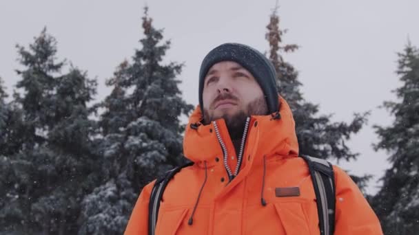 Una persona de 30 años en una chaqueta naranja brillante está mirando a la distancia con pinos en el fondo en un día de invierno nevado — Vídeos de Stock