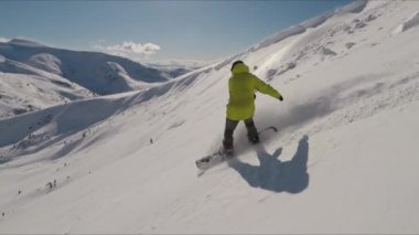 Bir snowboardcunun, dağda manzaralı bir şekilde gezerken havaya toz püskürttüğü bir fotoğraf.