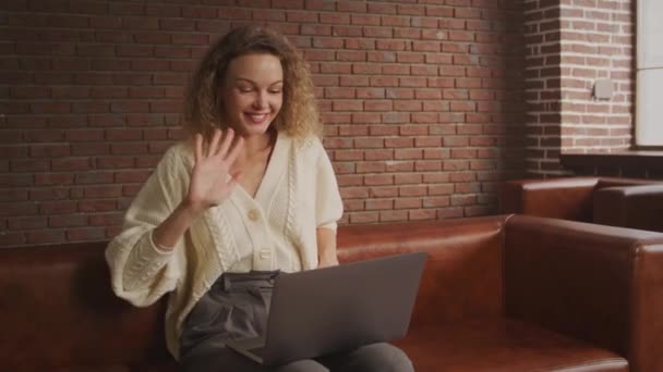 Kudrnaté blond dospělá žena sedí v pohovce s notebookem a mává rukou pozdravit své přátele na videohovor — Stock video
