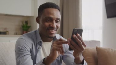 Yetişkin bir Afrikalı Amerikalı adamın kapalı alandaki siyah akıllı telefonuyla internette gezindiği bir sürü yüz ifadesi.