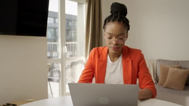 Jovem afro-americana bem sucedida mulher em uma jaqueta laranja brilhante trabalhando em um laptop em casa — Vídeo de Stock