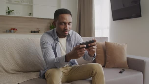 Африканський американець - дорослий дорослий чоловік грає в ігри на розумному телефоні і перемагає на дивані в своїй квартирі. — стокове відео