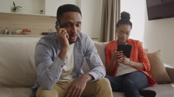 Dos jóvenes afroamericanos adultos se están enfriando en casa. Hombre hablando por teléfono y mujer navegando por la web en una tableta — Vídeos de Stock