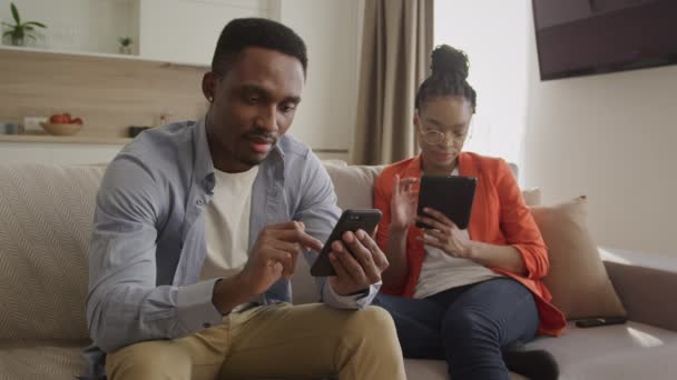 Zwei junge afrikanisch-amerikanische Erwachsene chillen zu Hause und surfen mit ihren Handys und Tablets im Internet — Stockvideo