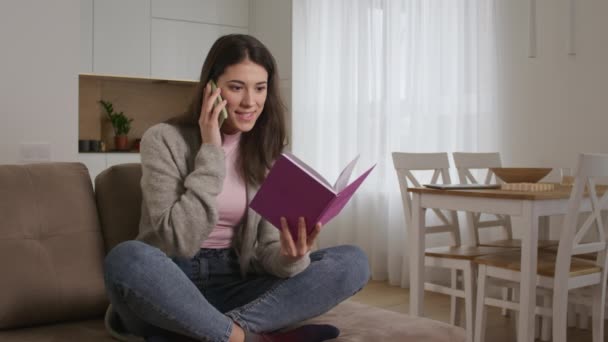 Mujer caucásica joven haciendo una conversación en un teléfono con un cuaderno rosa en sus manos en una sala de estar — Vídeos de Stock