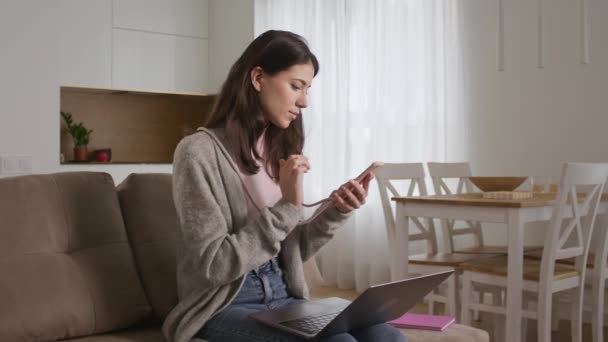 Glad ung 20-talet kvinna rulla på sin smartphone och byta till att bläddra på en bärbar dator i ett vardagsrum — Stockvideo