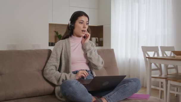 महिला उद्यमी एक उज्ज्वल अपार्टमेंट में घर पर अपने घुटनों पर लैपटॉप के साथ हेडफ़ोन का उपयोग करके ऑडियो बात कर रहा है — स्टॉक वीडियो