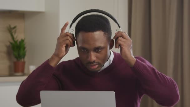 若いアフリカ系アメリカ人の男性は音楽でヘッドフォンを拾い、ラップトップの前でダンスを始めます — ストック動画