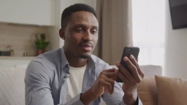 Oturma odasında akıllı telefonuyla yazı yazan Afro-Amerikan bir adamın portresi.