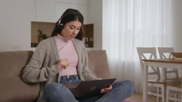 Jovem caucasiana cumprimentando seus amigos ou familiares em uma chamada de vídeo usando laptop e fones de ouvido — Vídeo de Stock