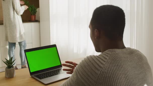 Pareja joven de raza mixta en su apartamento con un hombre afroamericano sentado frente a una computadora portátil de pantalla verde en una videollamada — Vídeo de stock
