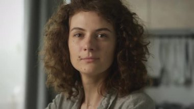 20 'li yaşlardaki kıvırcık beyaz kadın kameraya bakıyor ve oturma odasında gülümsüyor.