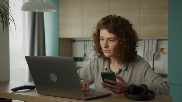 Jeune femme des années 20 naviguant sur son téléphone intelligent et double vérification sur son ordinateur portable tout en faisant des achats en ligne — Video