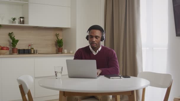 헤드폰을 끼고 노트북을 쓰고 있는 아프리카 계 미국인을 넓게 찍은 사진입니다. 먼 곳에서 일하는 콜 센터를 클로즈업하는 모습 — 비디오
