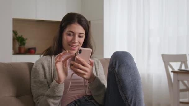 若い白人女性はウェブ上でソーシャルメディアを閲覧しながらスマートフォンで見たことを笑います — ストック動画