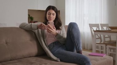 İnternette sörf yapan genç bir kadın oturma odasında akıllı telefonunu değişik yüz ifadeleriyle kullanıyor.
