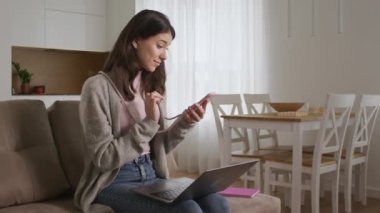 20 'li yaşlardaki mutlu bir kadın akıllı telefonunu kaydırıyor ve oturma odasında dizüstü bilgisayara geçiyor.