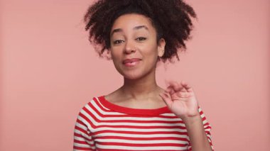 Neşeli kadın dostça el sallıyor, pembe stüdyo arka planında kameraya gülümsüyor..