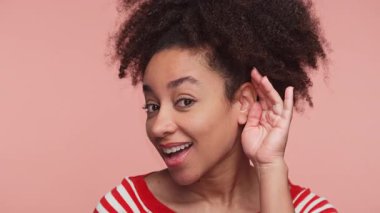 Gülümseyen genç bir kadın elini kulağına dayayıp, dinlediğini işaret ediyor ya da pembe arka planda birinin daha yüksek sesle konuşmasını istiyor..