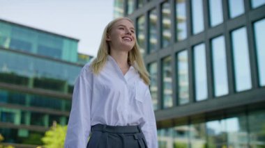 Resmi kıyafetli gülümseyen kadın şehir merkezinde dışarıda yürüyor, arka planda modern ofis binaları var..
