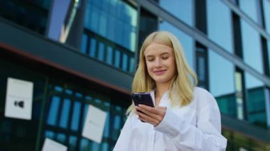 Şehirde yürürken akıllı telefon kullanan mutlu genç bir kadın. Yüzünde gülümsemeyle mesaj atıyor..