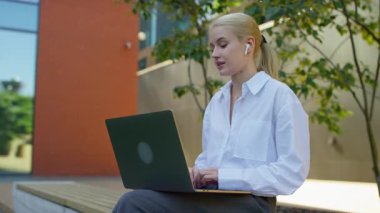 Modern bir binanın yanında oturan beyaz gömlekli genç bir kadın laptopuyla video görüşmesi yaparken konuşuyor..