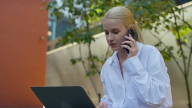 Beyaz gömlekli genç bir kadın dışarıda çalışıyor, bilgisayarını kullanırken telefonla konuşuyor, odaklanmış ve meşgul görünüyor..