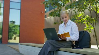 Dışarıda oturan genç bir kadın, dizüstü bilgisayarla çalışıyor ve sarı bir not defterine yazı yazıyor, ödevlerini tamamlamaya odaklanmış..