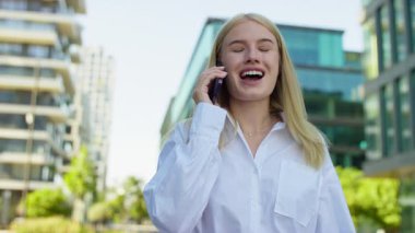 Neşeli sarışın kadın telefon görüşmesi sırasında gülüyordu. Etrafında modern binalarla güneşli bir caddede yürürken..