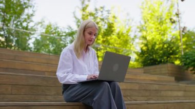 Dışarıdaki tahta basamaklarda oturan beyaz tişörtlü genç sarışın kadın, arka planda ağaçlarla dizüstü bilgisayarında yazmaya odaklanmış..