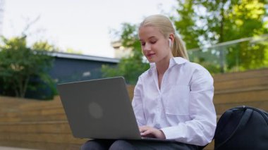 At kuyruğu ve beyaz gömlekli bir öğrenci dışarıda sırt çantasıyla otururken ve kulaklık takarken dizüstü bilgisayarda daktilo kullanıyor..