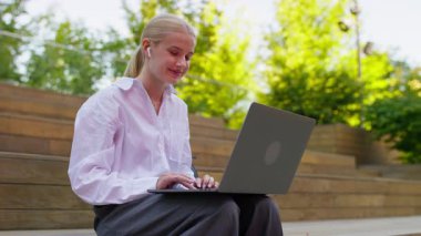 Beyaz gömlekli ve kulaklıklı bir öğrenci ahşap basamaklarda oturuyor, yeşil parktaki dizüstü bilgisayarının içeriğini inceliyor..