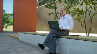 Beyaz gömlekli ve kulaklıklı genç bir kadın dizüstü bilgisayarla bankta oturuyor ve modern kampüs binalarının yakınındaki online bir sınıfta aktif olarak çalışıyor..