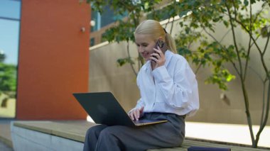 Beyaz gömlekli genç bir kadın bankın dışında oturuyor, dizüstü bilgisayar kullanırken telefonla konuşuyor, yakınlarda defterler ve ağaçlar var..