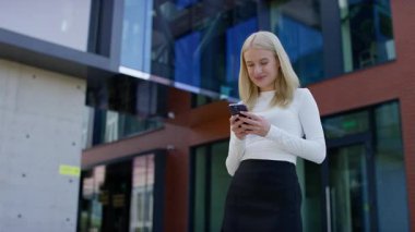 Güneşli bir günde modern bir ofis binasının önünde dikilirken beyaz kollu, üst üste mesajlaşan sarışın bir kadın..