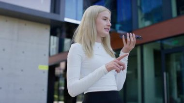 Beyaz bluzlu sarışın kadın ağzının önünde akıllı telefon tutuyor ve modern cam binanın yakınında bir sesli mesaj kaydediyor..