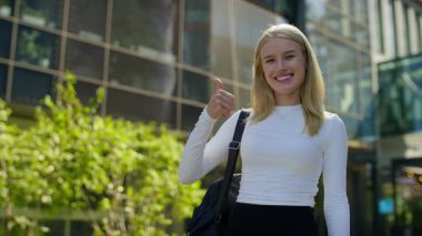 Beyaz bluzlu gülümseyen sarışın kadın güneşli bir günde modern cam bir binanın önünde dikilirken baş parmak hareketi yapıyor..