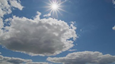 Parlak güneş bulutların arasından mavi bir gökyüzünde parlıyor. Bulut hareketinin hızlandırılması ve değişen ışık desenleri için ideal..