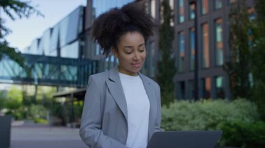 Dışardaki bankta oturan ceketli, gülümseyen bir kadın, modern kentsel alanda yeşil bir dizüstü bilgisayar kullanıyor..