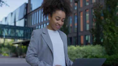 Şehir ortamında spor ceketli, gülümseyen bir kadın. Arkasında yeşillik olan bir dizüstü bilgisayarda kayıyor ya da geziniyor..