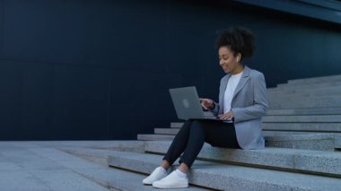 Genç profesyonel kadın şehir basamaklarında oturuyor, gülümsüyor ve dışarıda dizüstü bilgisayarla çalışırken kablosuz kulaklık takıyor..