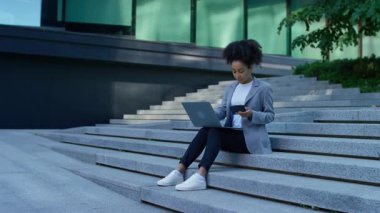 Modern şehir ortamında merdivenlerde oturan genç profesyonel bir kadın, finans veya bütçe yönetimi için dizüstü bilgisayar ve hesap makinesi kullanıyor.