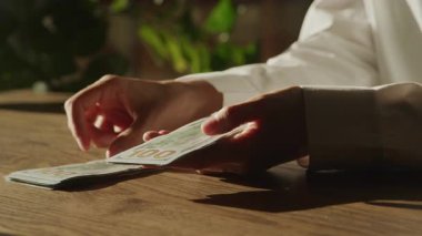 Eller beyaz gömlekli, ahşap bir masada 100 'lük banknotları dikkatlice ayır, arka planda güneş ışığı ve yeşillik olsun..