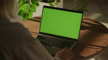 Kullanıcı, ahşap bir masanın üzerinde yeşil ekranlı, kapalı bitkiler ve yumuşak doğal ışıklandırmayla çevrili bir dizüstü bilgisayarla etkileşir..