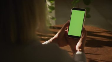 Elinde yeşil renkli bir ekran olan akıllı telefonu olan, tahta bir masada oturan ve doğal güneş ışığı alan bir kişinin arka görüntüsü..