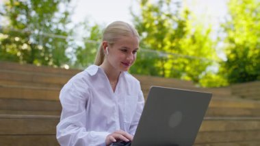 Beyaz tişörtlü genç bir kadın güneşli bir ortamda kablosuz kulaklık takarken dizüstü bilgisayar çalışıyor ya da çalışıyor..