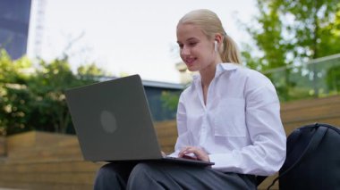 Sarışın kadın, kablosuz kulaklık takıyor ve beyaz gömlek, gün ışığında dışarıda, tahta merdivenlerde otururken dizüstü bilgisayarda kayıyor..