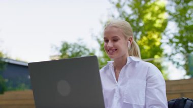 Kulaklıklı gülümseyen kadın, güneşli bir günde internette dolaşırken laptop ekranına bakıyor..