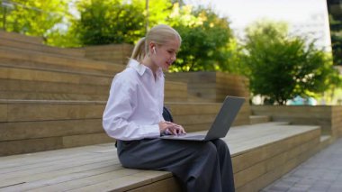 Kulaklık takan şaşırmış ve heyecanlı bir kadın güneşli bir ortamda dizüstü bilgisayarının ekranına bakarken sevinçle tepki veriyor..